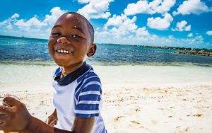 A smiling child plays on a white sandy beach near blue waters.