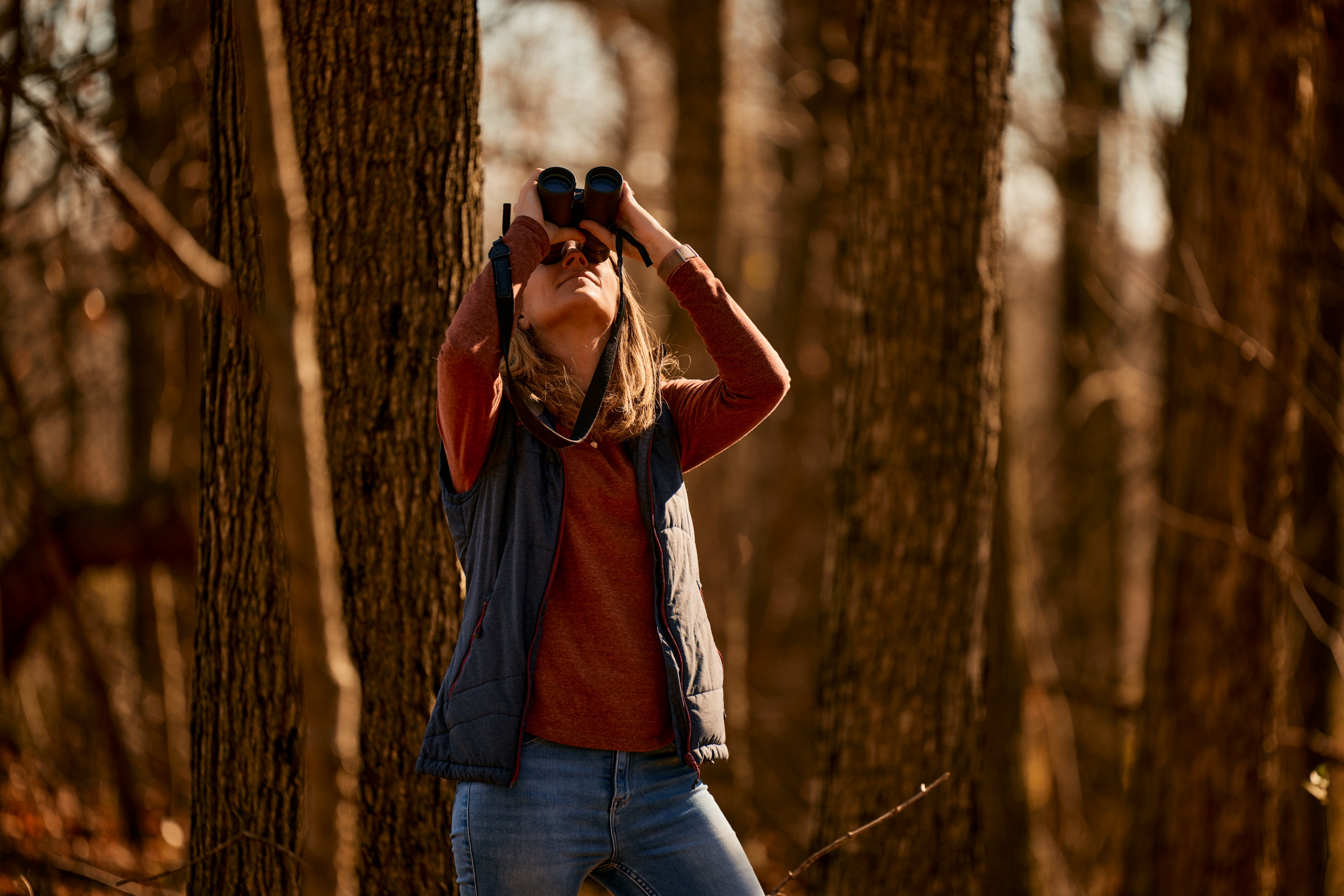 A woman in a forest looking up through binoculars. 