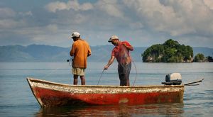 Two fishermen stand in a red boat, pulling nets along the water. 