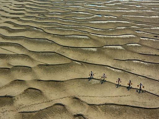 Aerial view of five people carrying water on their shoulders across a desert landscape.